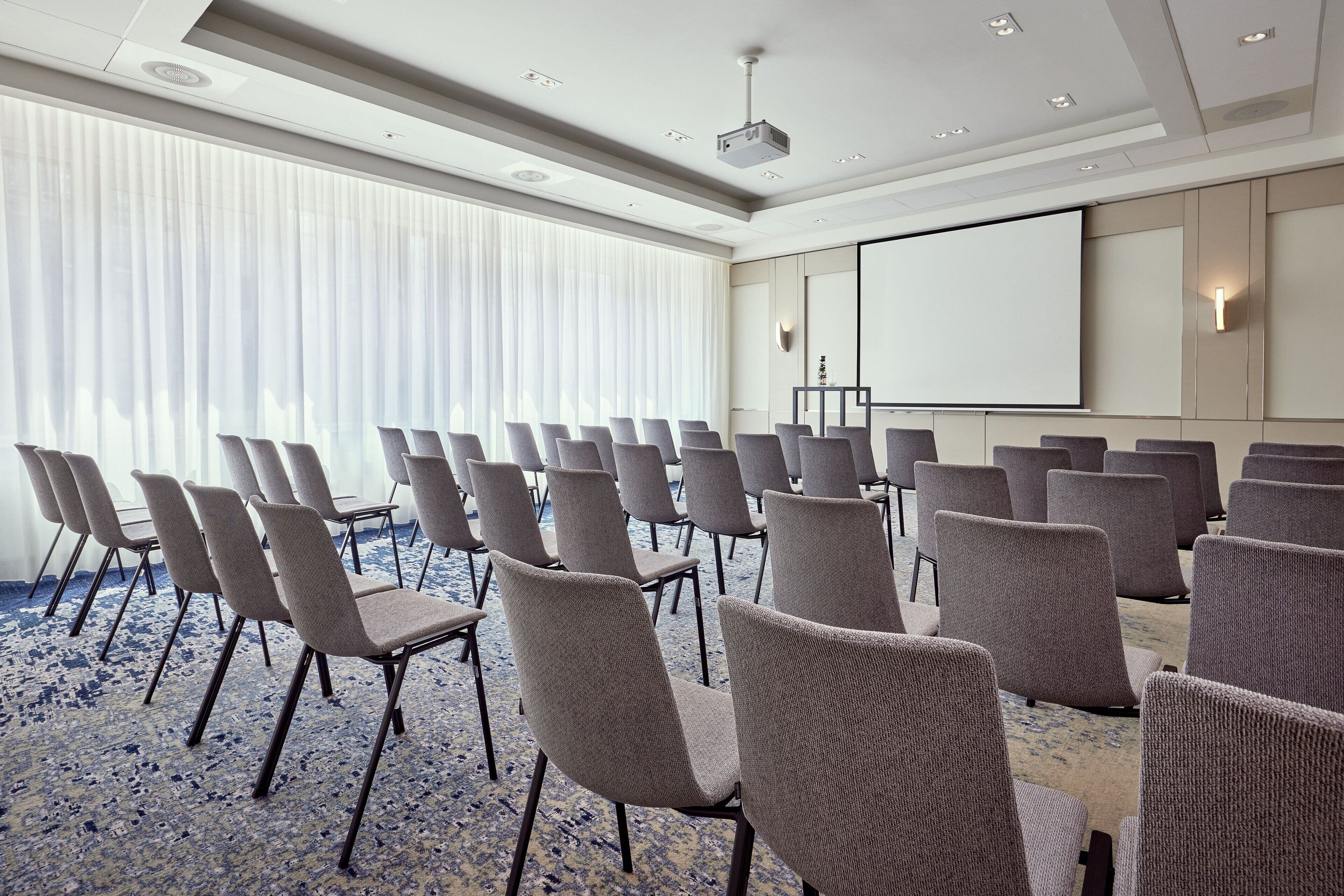 Una sala conferenze con file di sedie, uno schermo per proiezioni e un'ampia finestra con persiane.