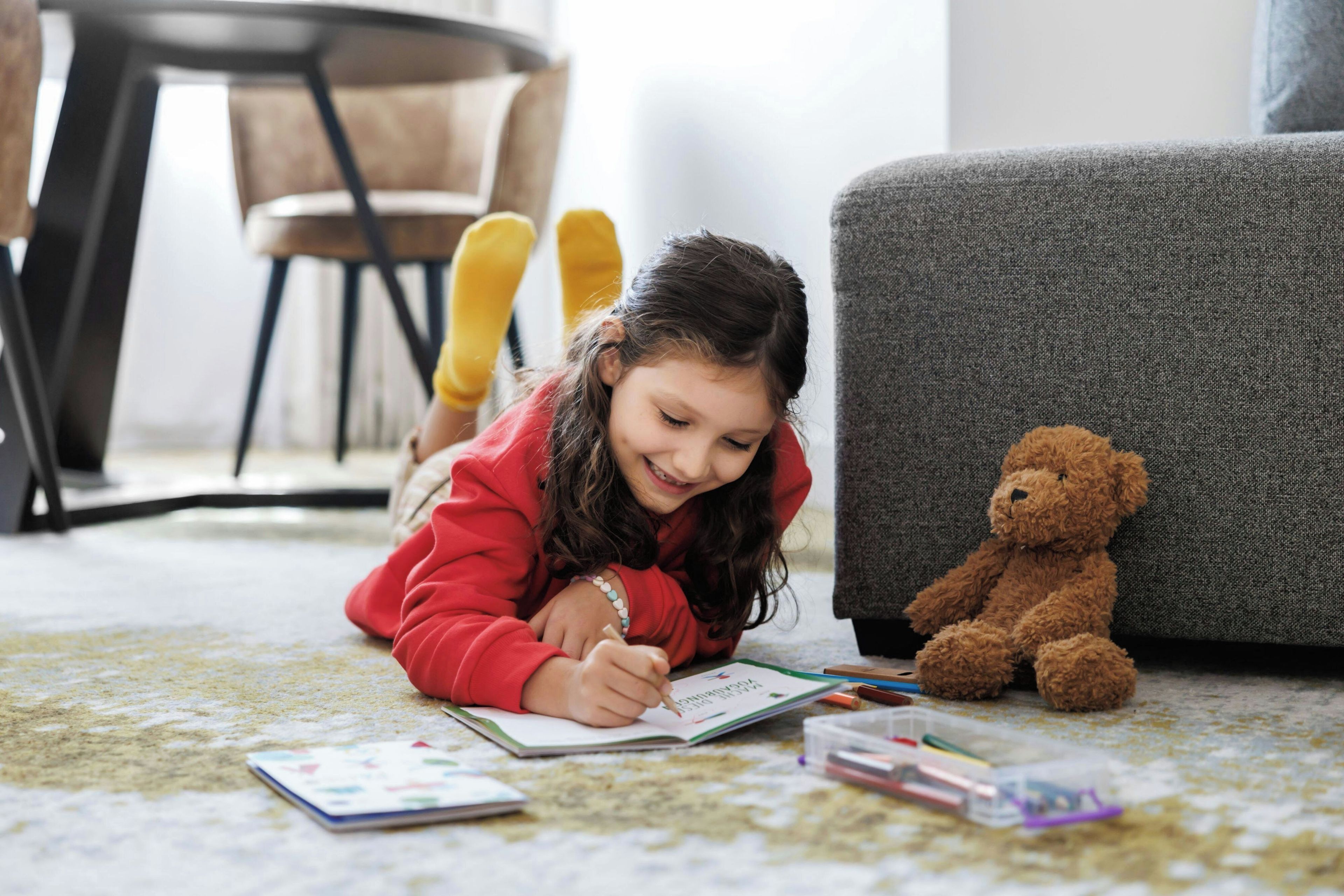 A child writing at a table with a teddy bear beside them, a chair, and a sofa in the background.