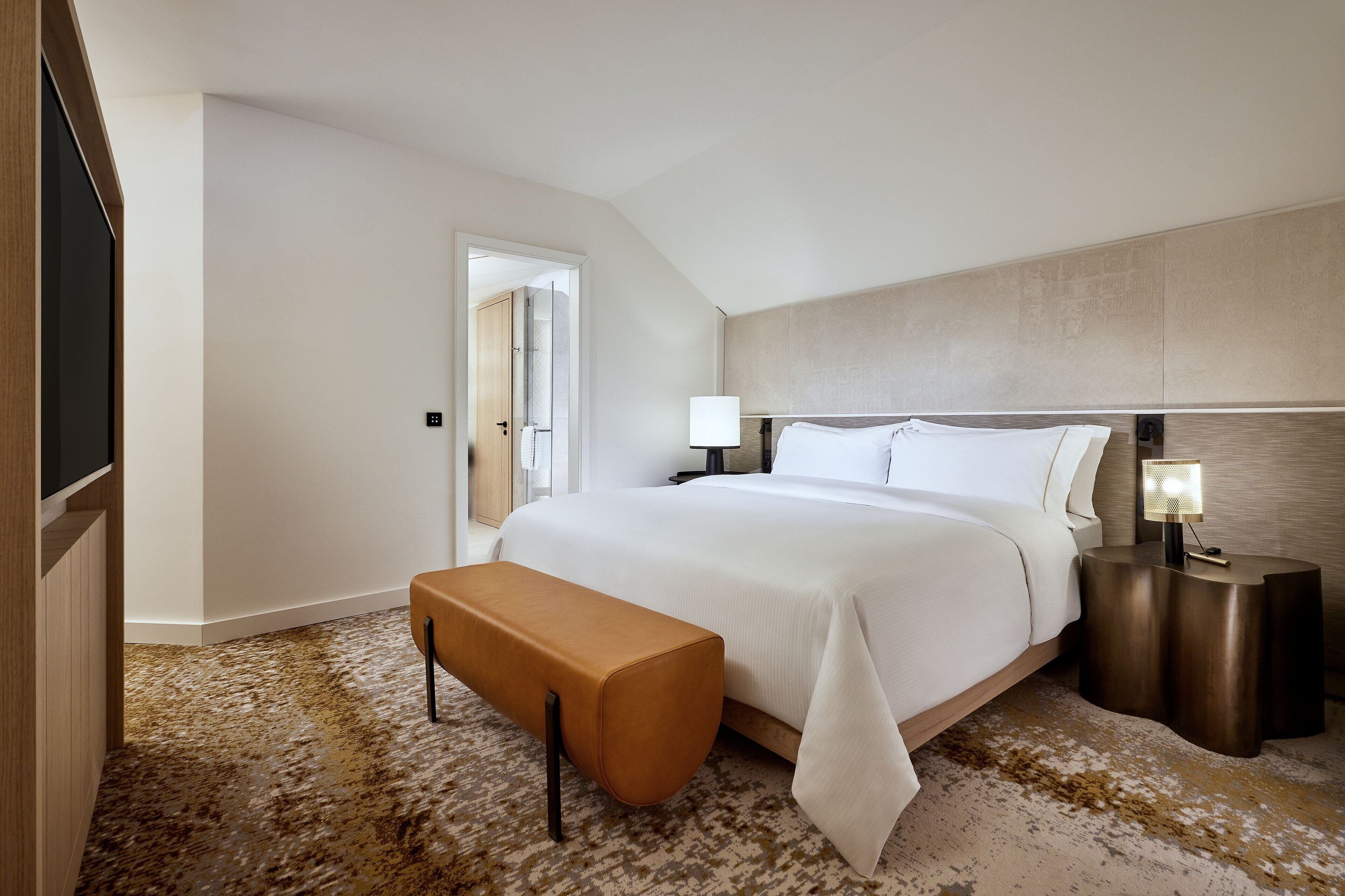 A modern hotel room with a large bed, a bench, two bedside tables with lamps, and a door leading to another room.