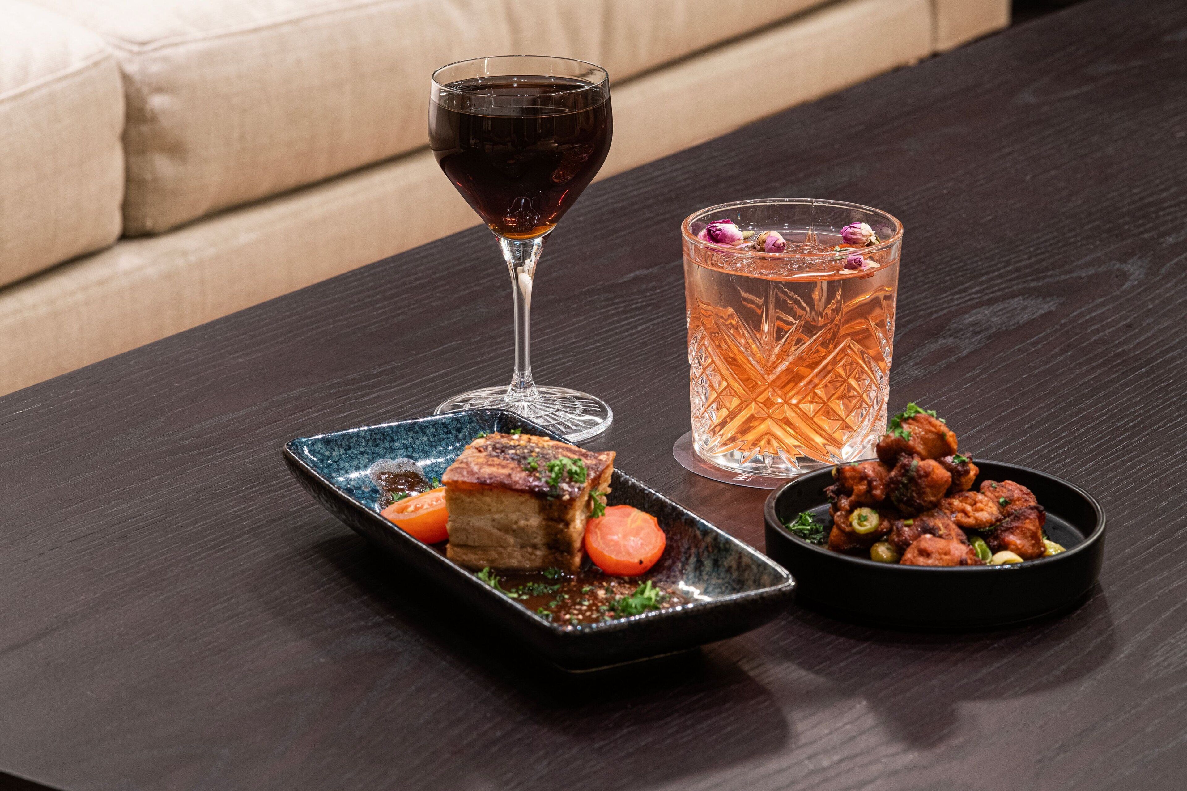 A dark wooden table with a plate of food, a glass of red wine, and a glass of iced tea with a floral garnish.