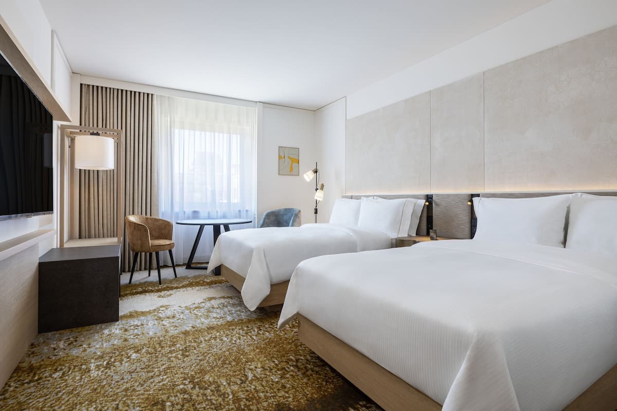 A hotel room with two beds, a desk, a chair, and a large window with curtains.