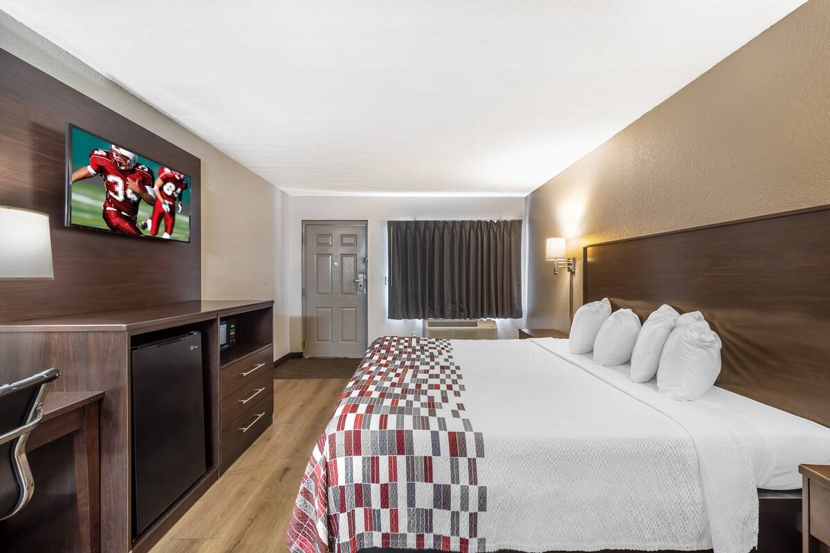 A hotel room with a bed, a TV mounted on the wall, a desk with a chair, and a window with curtains.