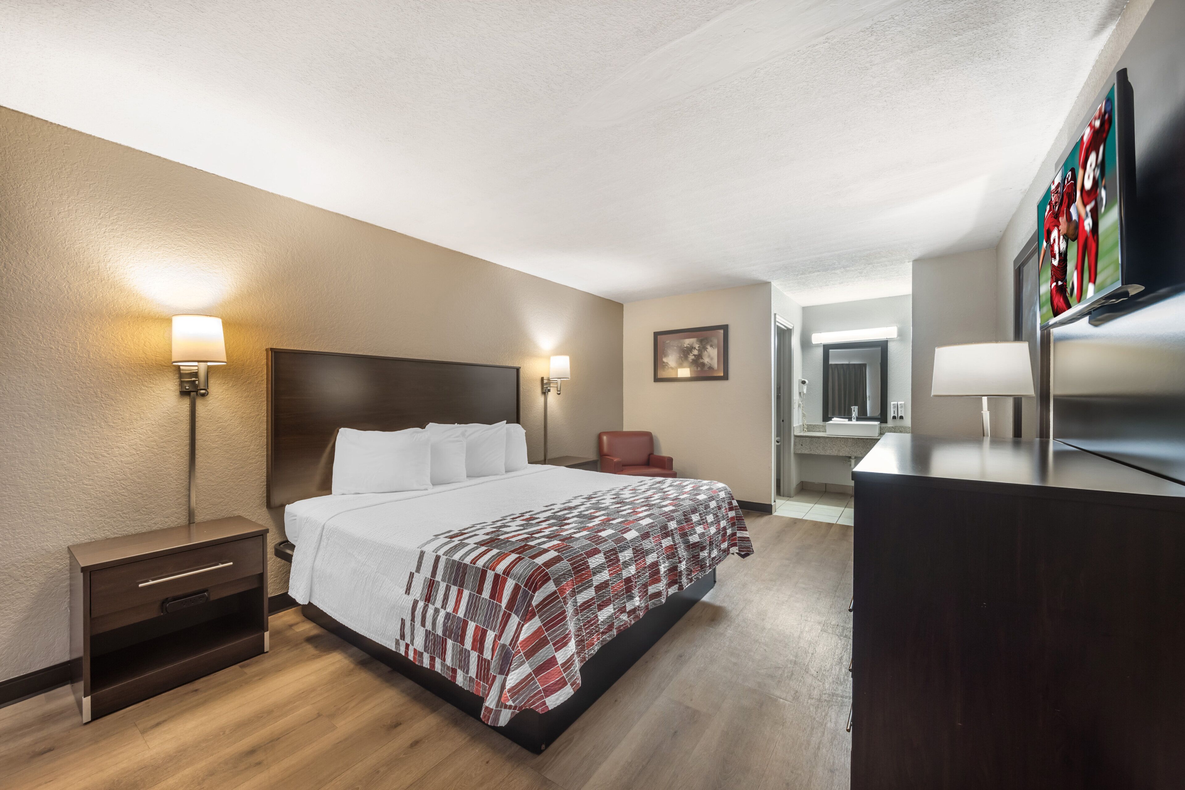 A hotel room with a large bed, bedside lamps, a desk with a chair, a television, and a framed picture on the wall.