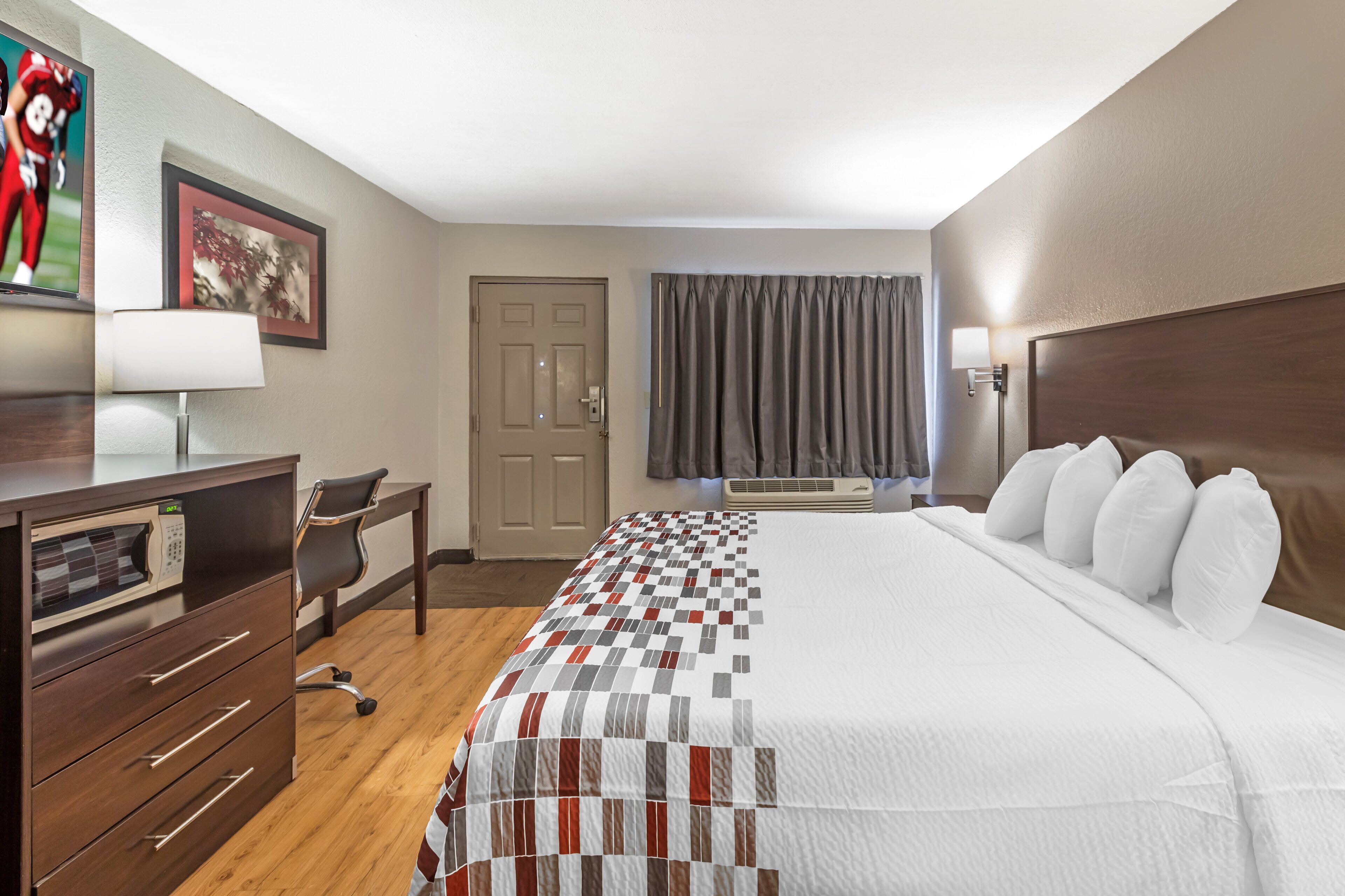 A hotel room with a bed, a desk, a chair, a microwave, a door, and a window with curtains.
