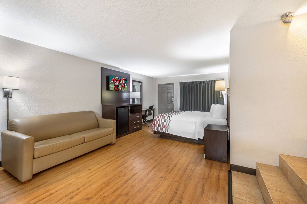 A hotel room with a wooden floor, a beige sofa, a bed with a checkered bedspread, a desk with a TV, and a door with curtains.
