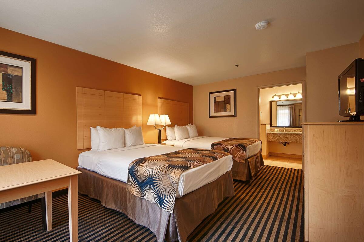 A hotel room with two beds, a desk, a chair, a framed artwork, a mirror, and a television.