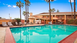 A swimming pool with lounge chairs and umbrellas, surrounded by palm trees and a building with balconies.
