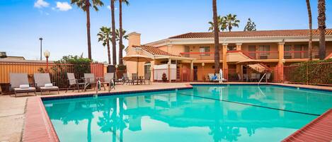 A swimming pool with lounge chairs and umbrellas, surrounded by palm trees and a building with balconies.