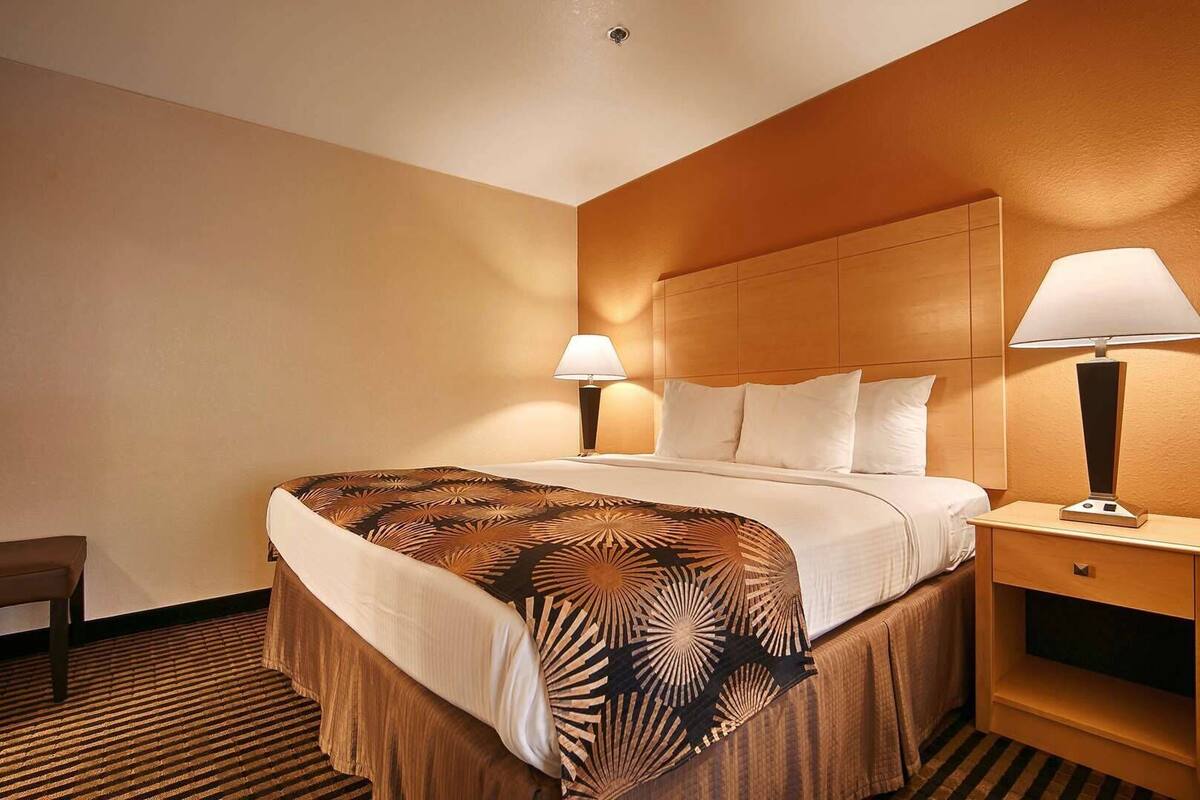 A hotel room with a bed, two bedside lamps, a wooden nightstand, a chair, and a patterned bedspread.