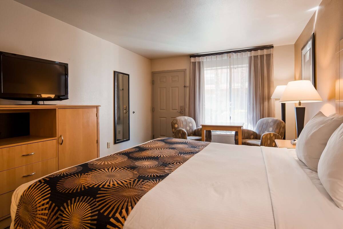 A hotel room with a bed, a TV, a desk, two chairs, a lamp, and a window with curtains.