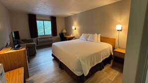 Ein Hotelzimmer mit einem großen Bett, einem Schreibtisch mit Computer, einem Sessel, einem Fenster mit Vorhängen und zwei Nachttischlampen.