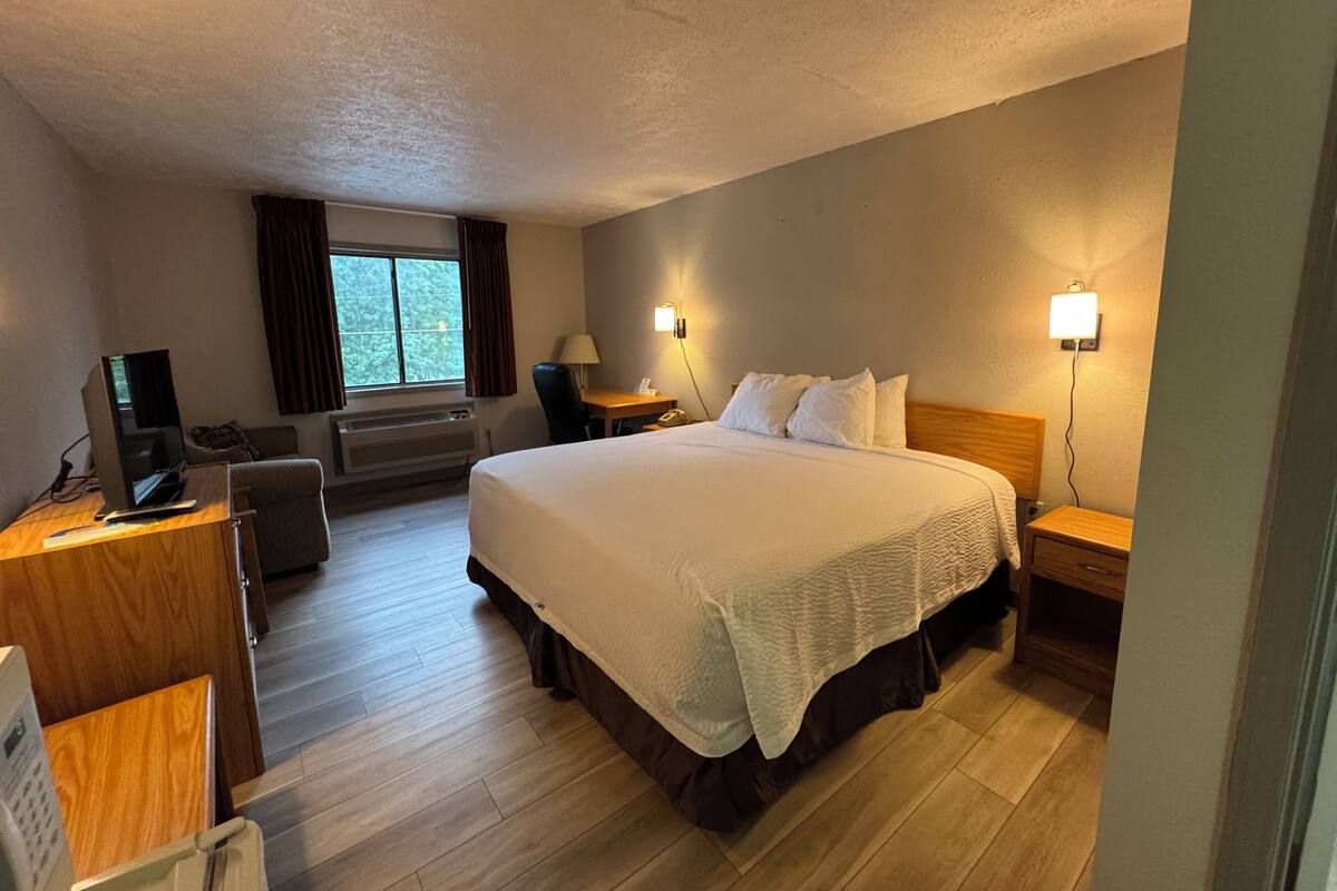 A hotel room with a large bed, a desk with a computer, a chair, a window with curtains, and two bedside lamps.