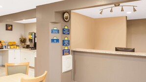 Eine Hotelhalle mit einer Rezeption, einem Check-in-Schalter, einer Sitzecke mit Tisch und Stühlen sowie einer kleinen Küchenzeile im Hintergrund.