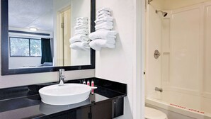 Une salle de bain avec un lavabo blanc, un plan de toilette noir et un miroir au-dessus. On y trouve un porte-serviettes avec des serviettes pliées et un espace douche avec une baignoire blanche.