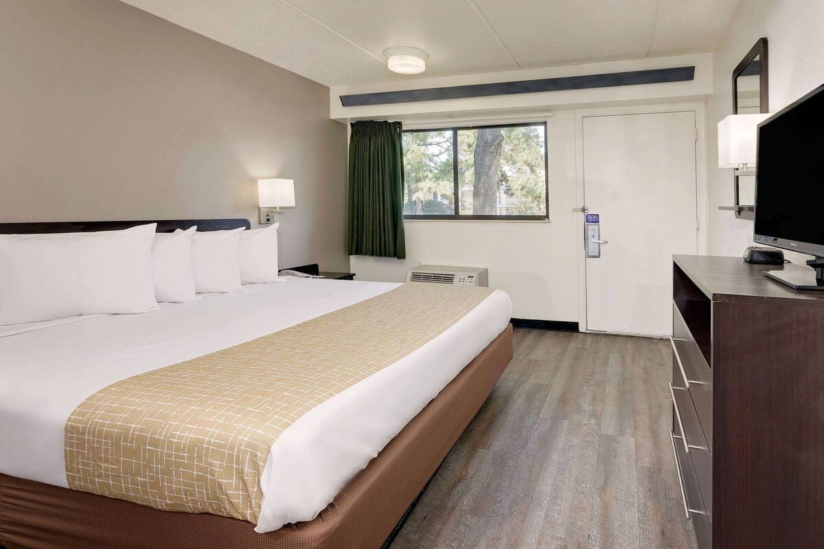 Ein Hotelzimmer mit einem großen Bett, einem Schreibtisch mit Fernseher, einem Fenster mit Vorhängen und einer Tür mit Schloss.
