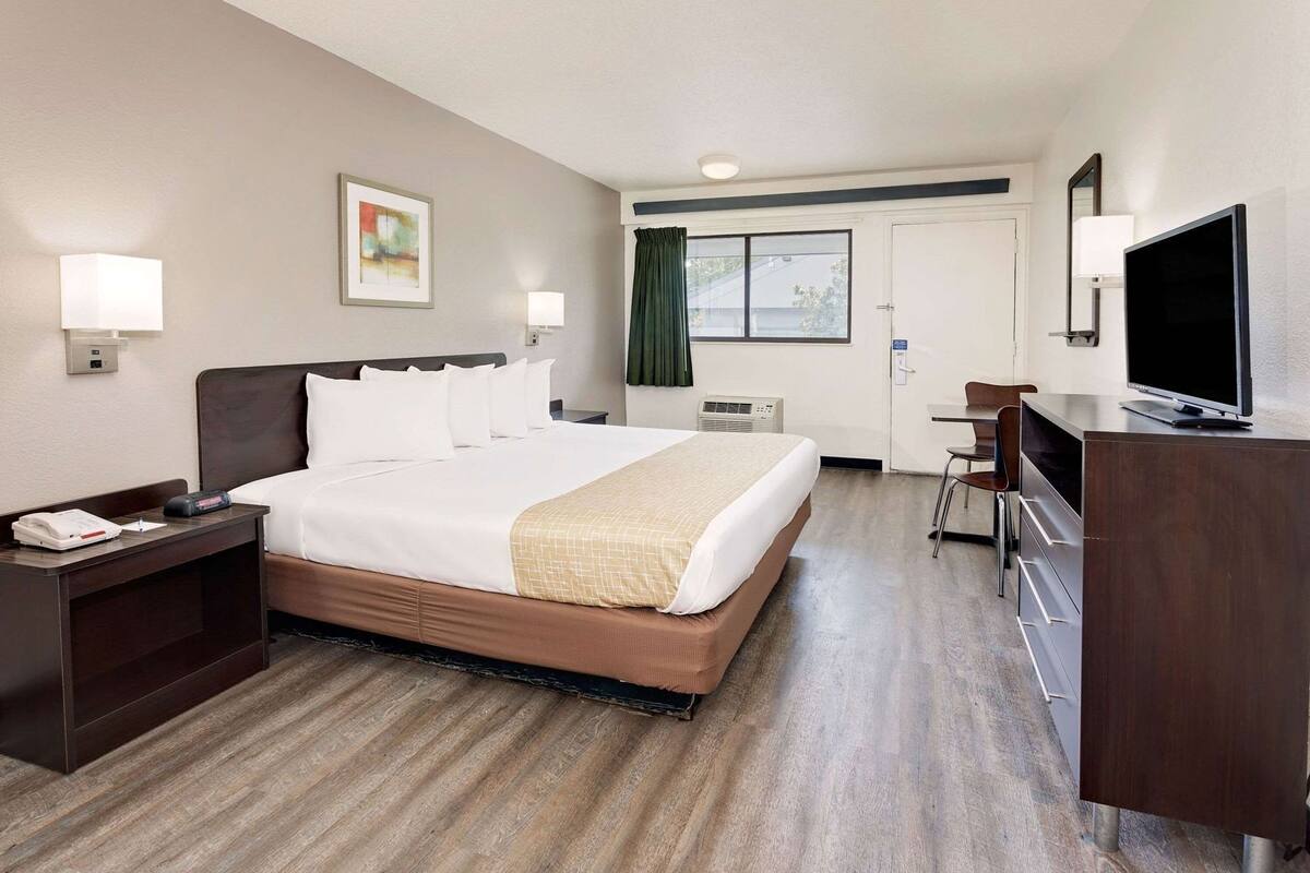 Ein Hotelzimmer mit Bett, Nachttisch, Schreibtisch, Stuhl, Fernseher und einem Fenster mit Vorhängen.