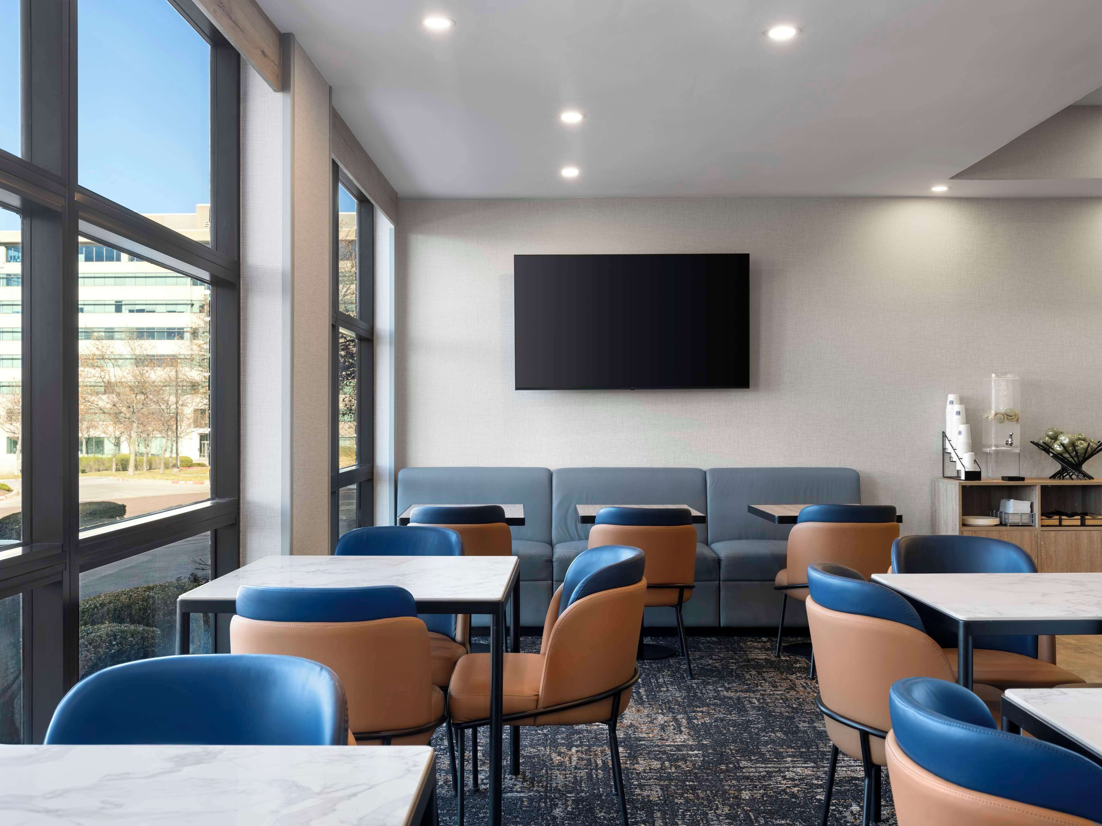A modern hotel room with large windows, a flat-screen TV, a sofa, and a dining area with tables and chairs.