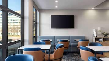 A modern hotel room with large windows, a flat-screen TV, a sofa, and a dining area with tables and chairs.