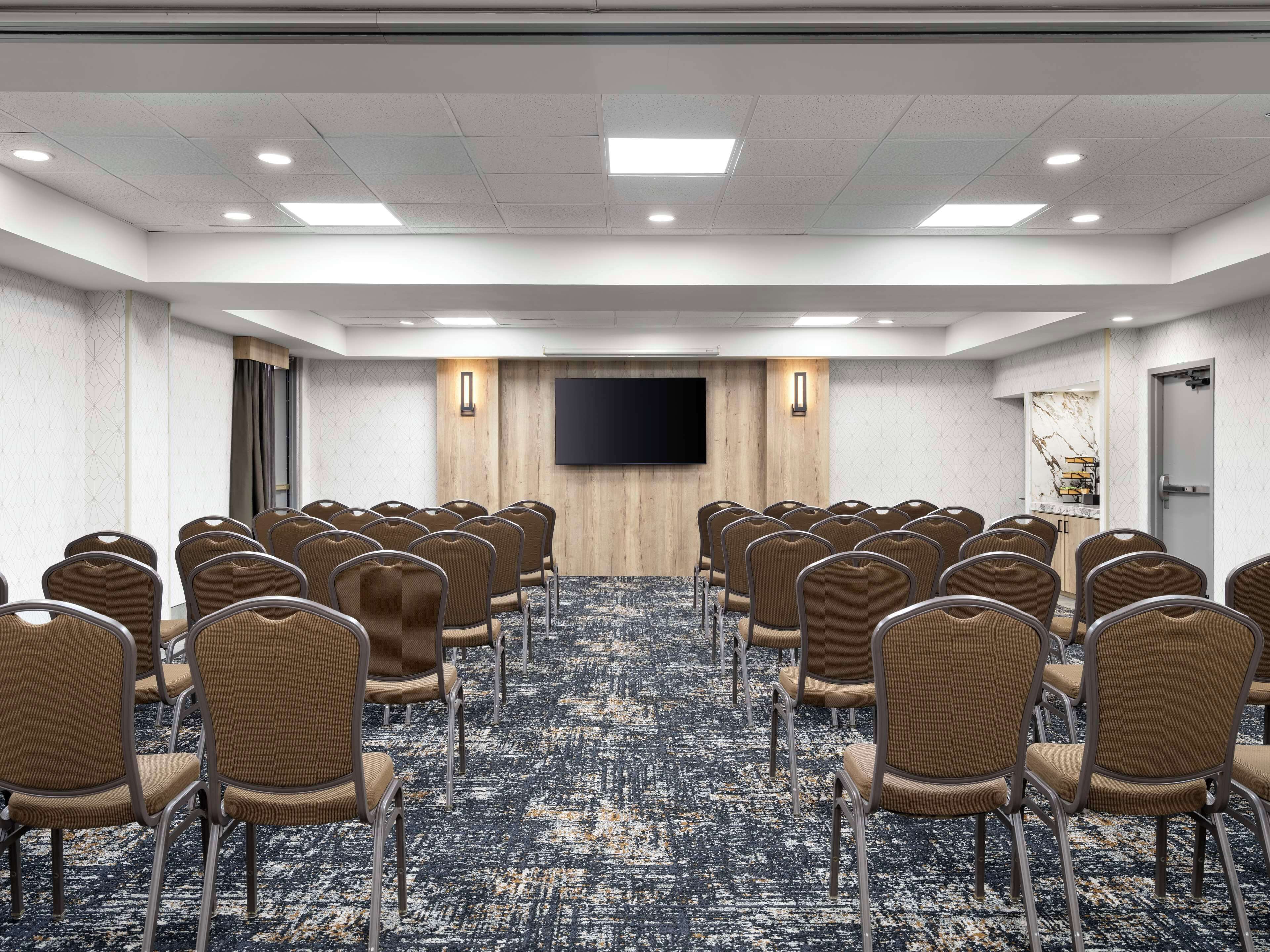 Un salón de conferencias con filas de sillas, un televisor de pantalla plana grande en la pared y una alfombra estampada.
