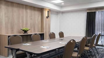 Un salón de conferencias con una mesa larga, seis sillas, una planta pequeña en una mesita auxiliar y una ventana con cortinas.