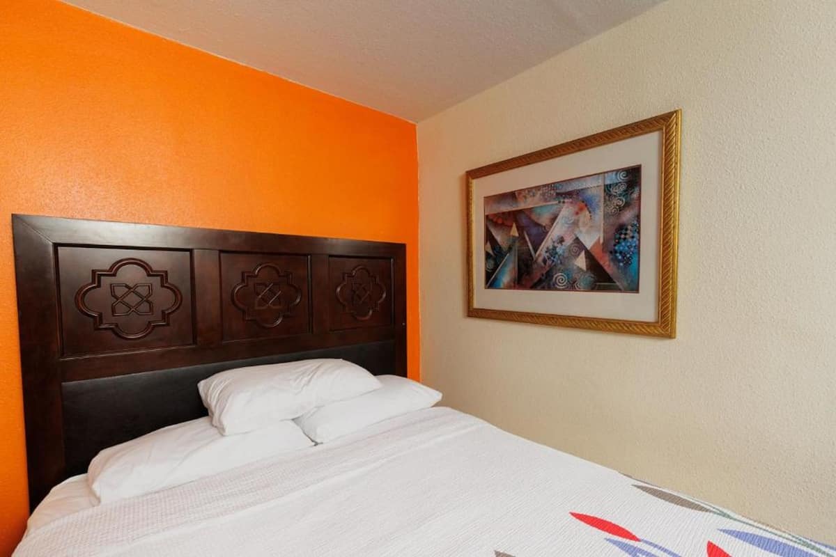 Une chambre d’hôtel avec un lit, un tableau abstrait encadré et une tête de lit en bois ornée de motifs décoratifs.