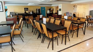 Ein Hotelrestaurant mit mehreren Tischen und Stühlen, eine Bar mit Kühlschrank und Kaffeemaschine, sowie eine blaue Tür im Hintergrund.