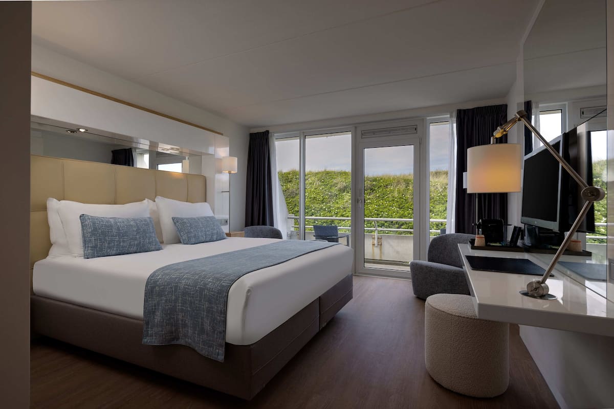 A modern hotel room with a large bed, a desk with a computer, and a view of the outdoors.
