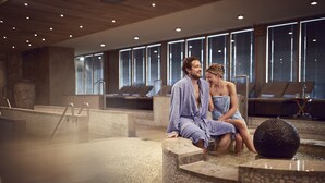 A couple in bathrobes sitting by a stone fountain in a spa setting with large windows and lounge chairs in the background. - WestCord Strandhotel Seeduyn (Vlieland)