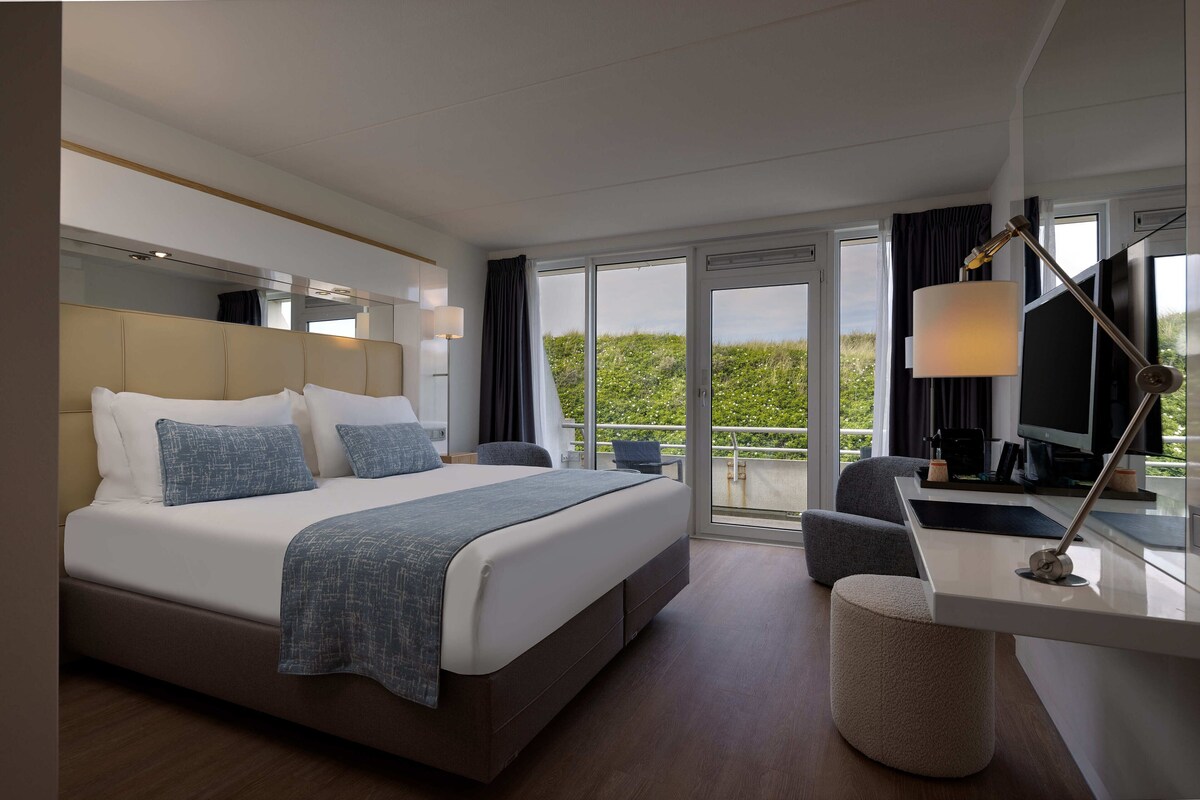 A modern hotel room with a large bed, a desk, a chair, and a view of greenery outside.