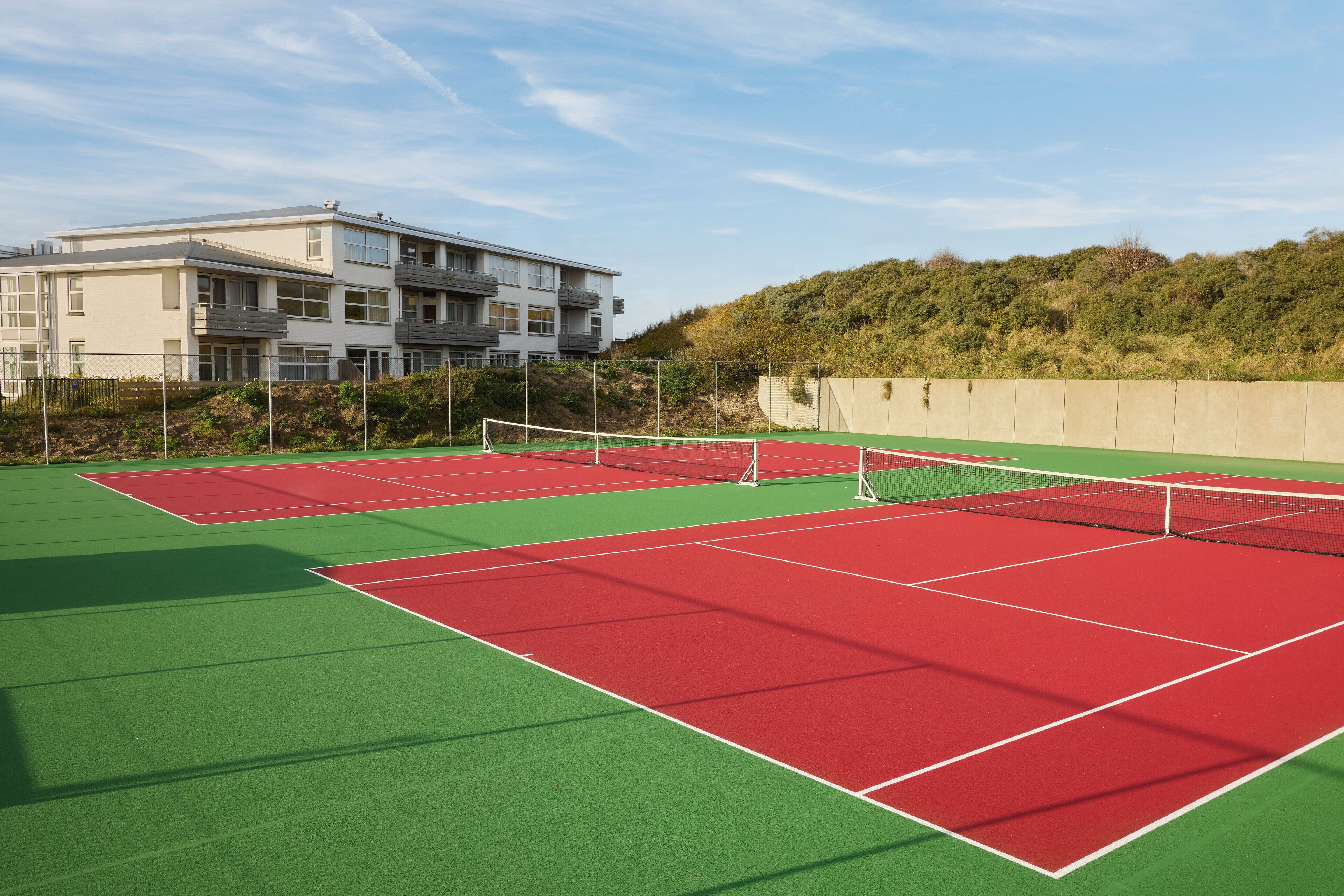A tennis court with a net, surrounded by a fence, and a building in the background.