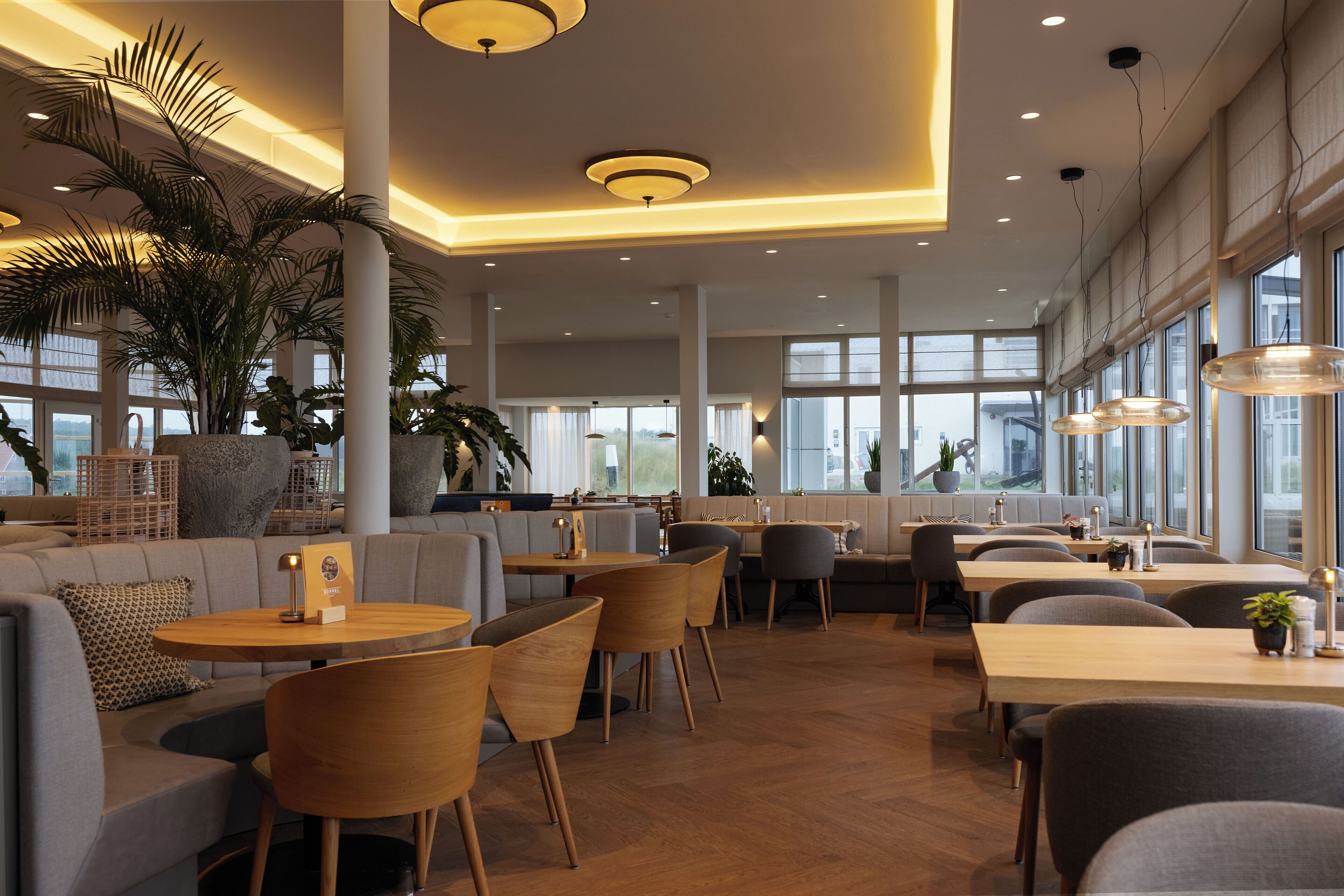 A modern restaurant interior with high ceilings, large windows, and a mix of seating options including tables and sofas.
