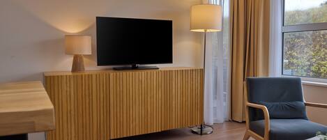 Ett modernt hotellrum med ett bord i trä, en platt-TV, en lampa, en stol och ett fönster med gardiner.