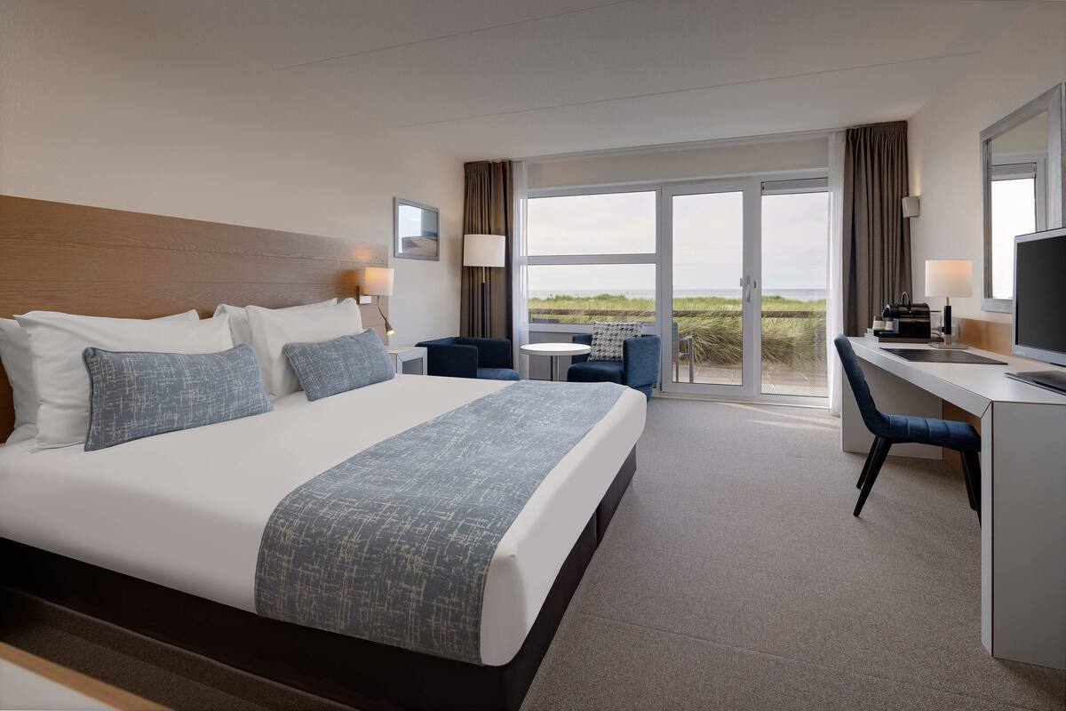 A hotel room with a large bed, a desk, a chair, and a view of the beach through the glass door.