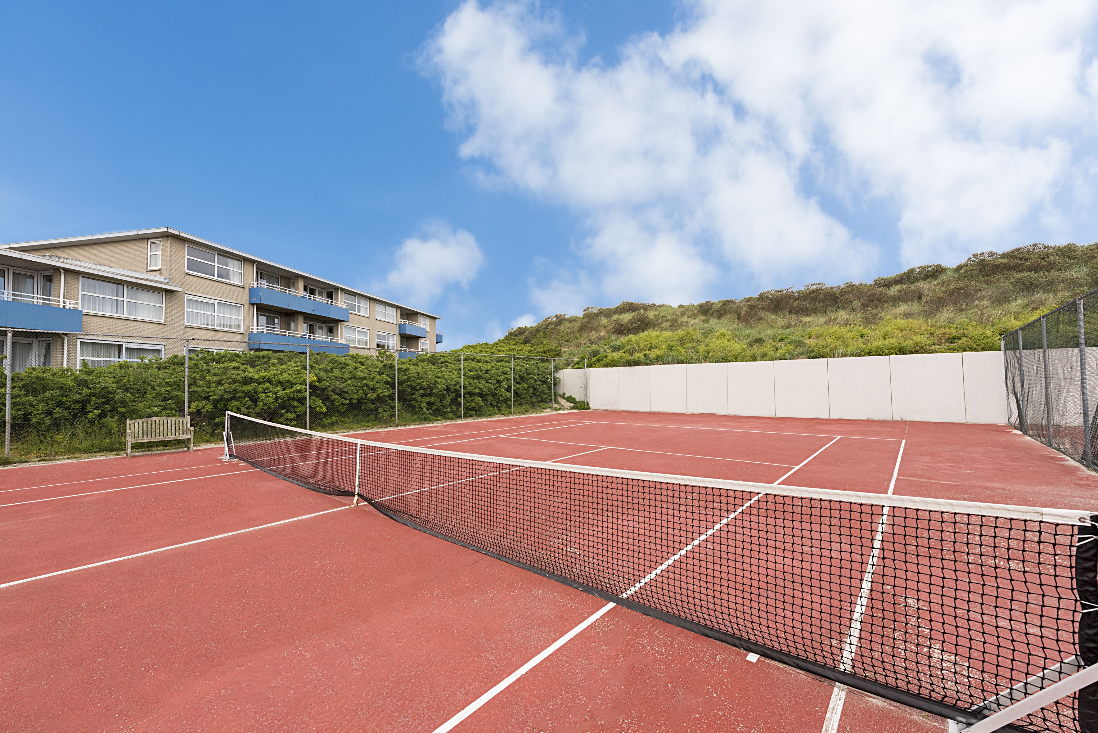 A tennis court with a net, surrounded by a fence and a building with balconies.