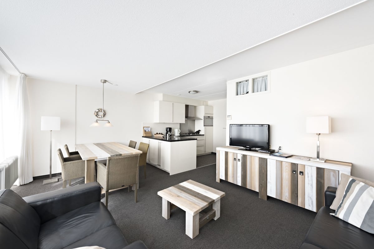 A modern hotel room with a living area, dining space, kitchenette, and a flat-screen TV.