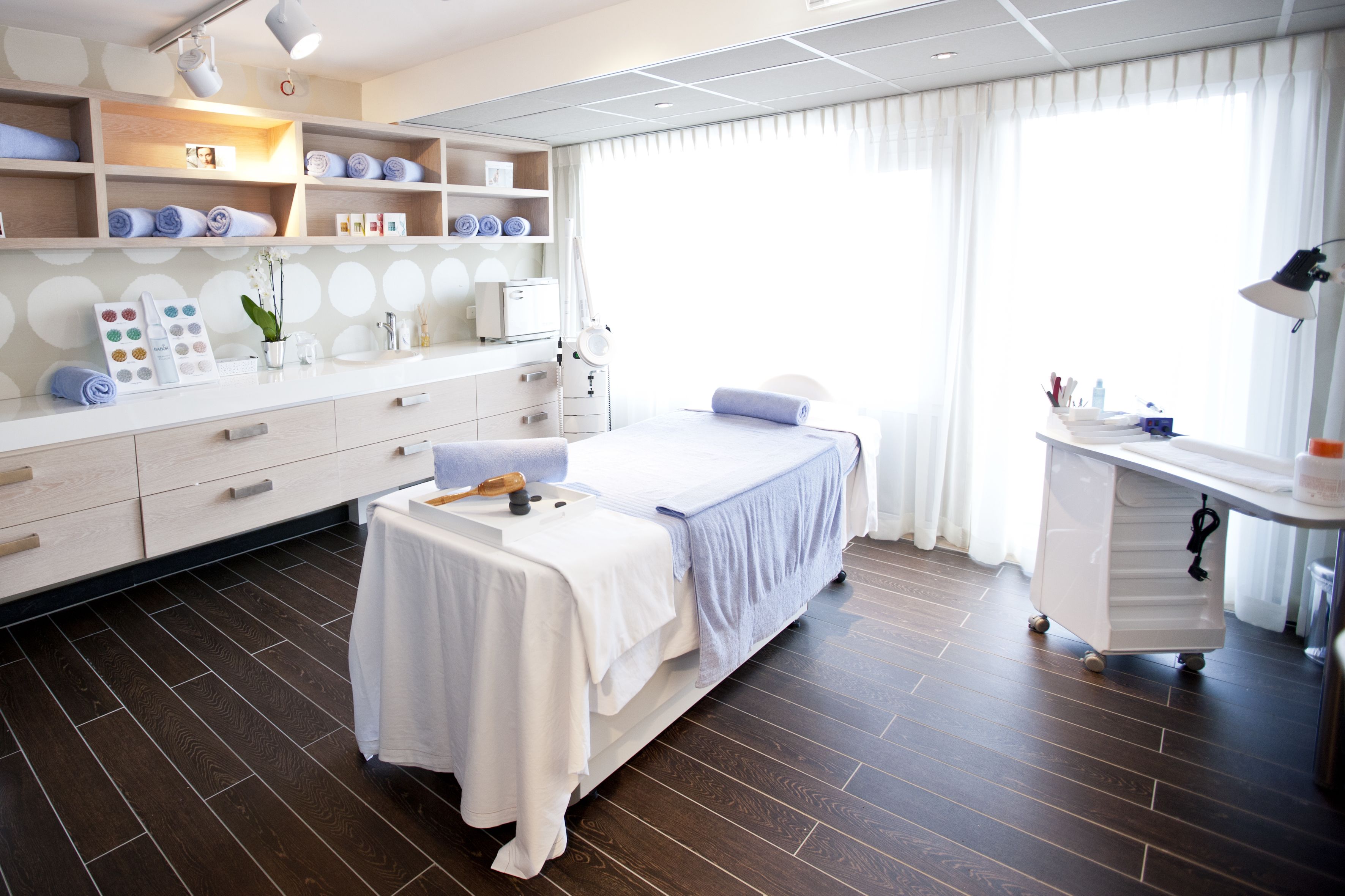 A modern spa room with a bed, a massage table, a vanity with a mirror, shelves with towels, and a large window with curtains.