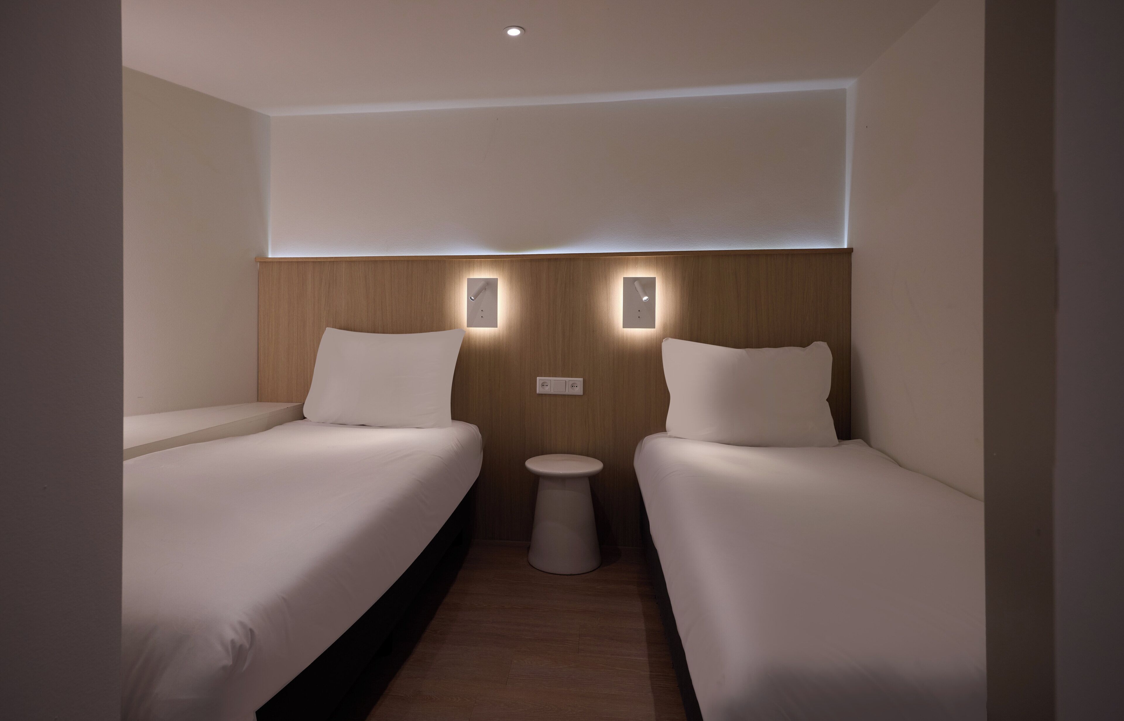 A hotel room with two single beds, a small round table, and wall-mounted lighting.