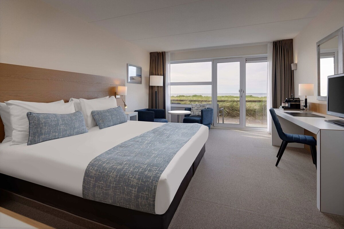 A hotel room with a large bed, a desk, a chair, a TV, and a view of the beach.