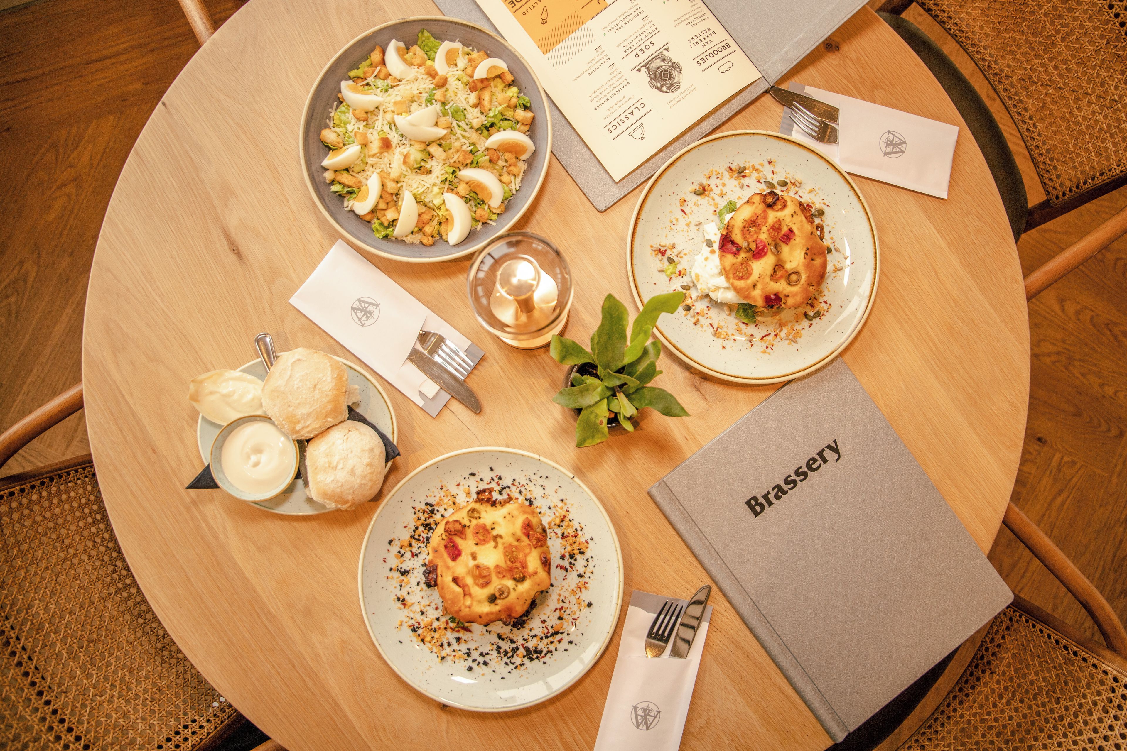 Une table ronde en bois, avec un menu, des assiettes de nourriture, un petit panier de pain et une bougie.