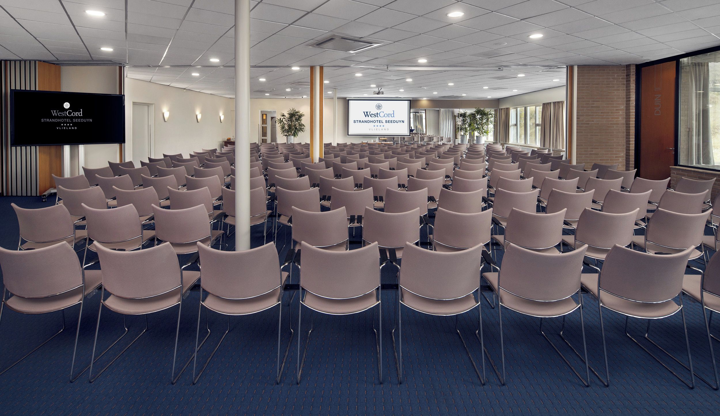 Sala de conferências com fileiras de cadeiras, uma tela grande exibindo a marca WestCord e uma parede de tijolos.
