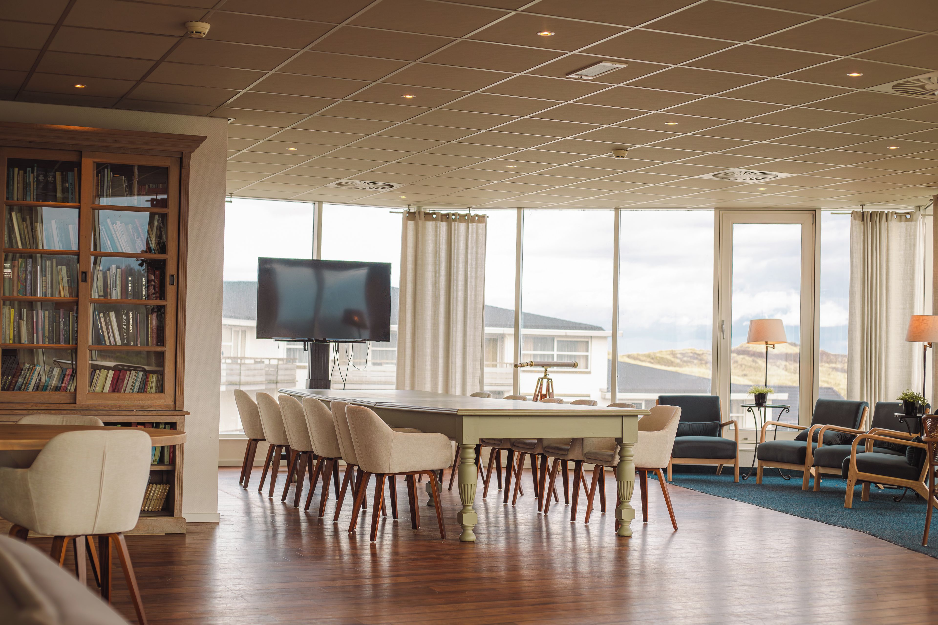 A spacious room with a large dining table, multiple chairs, a television, a bookshelf, and a view of the outdoors.