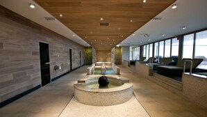 A modern indoor pool area with a central water feature, seating along the sides, and a wooden ceiling. - WestCord Strandhotel Seeduyn (Vlieland)