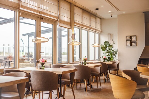 A restaurant interior with large windows, wooden tables, and chairs. - WestCord Strandhotel Seeduyn (Vlieland)