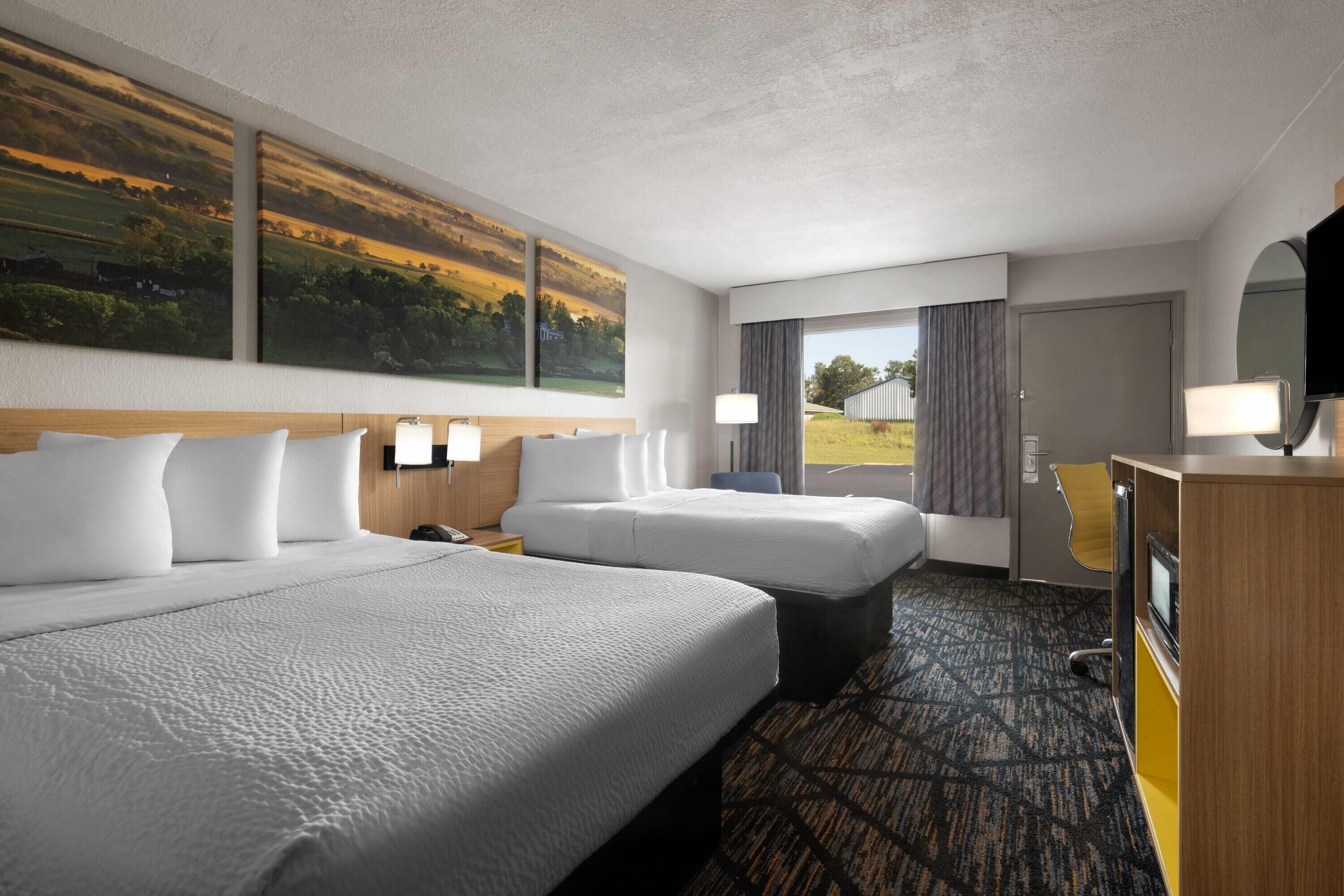 A hotel room with two beds, a desk, a TV, and a large window with a view of a field.