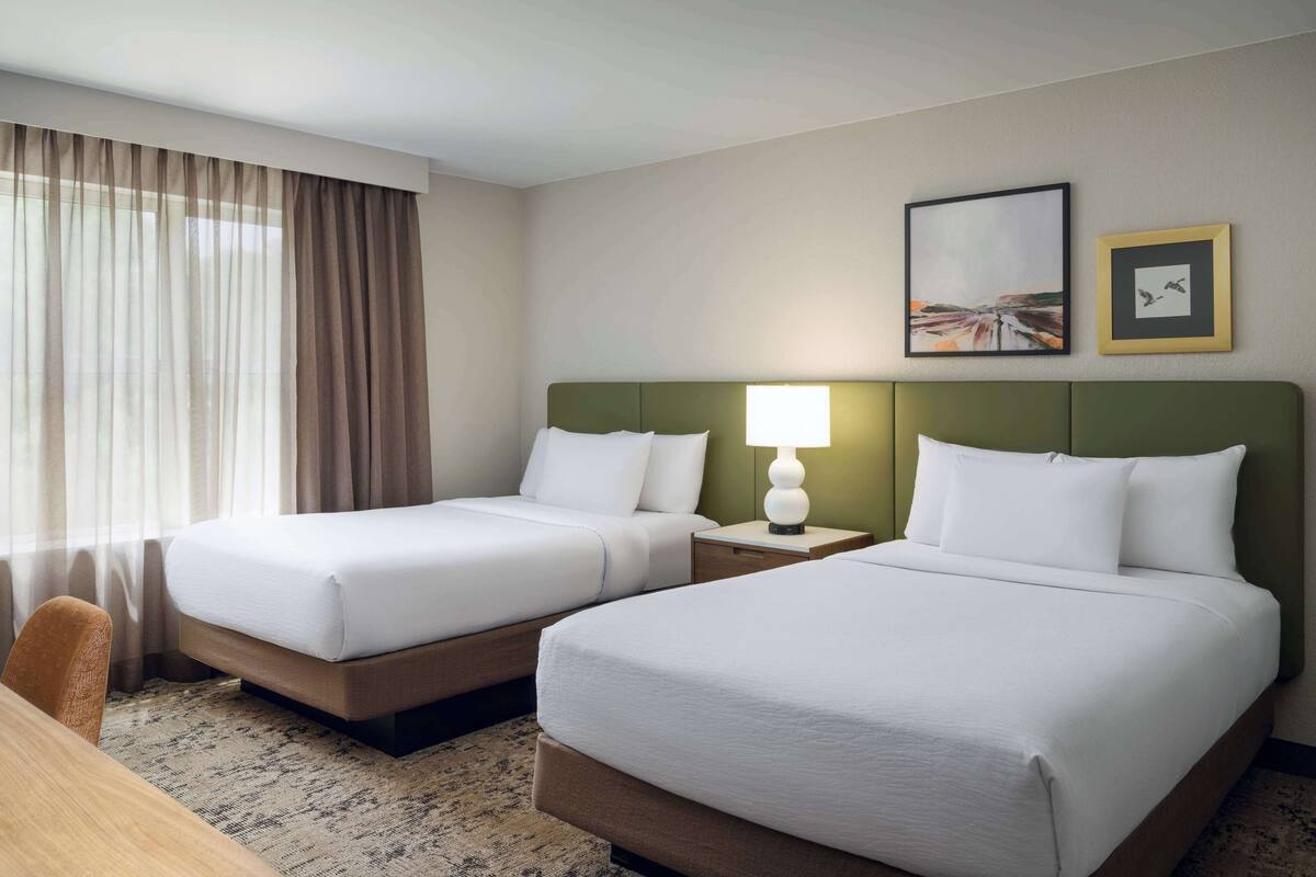 A hotel room with two single beds, a wooden desk, a chair, a lamp, and two framed pictures on the wall.