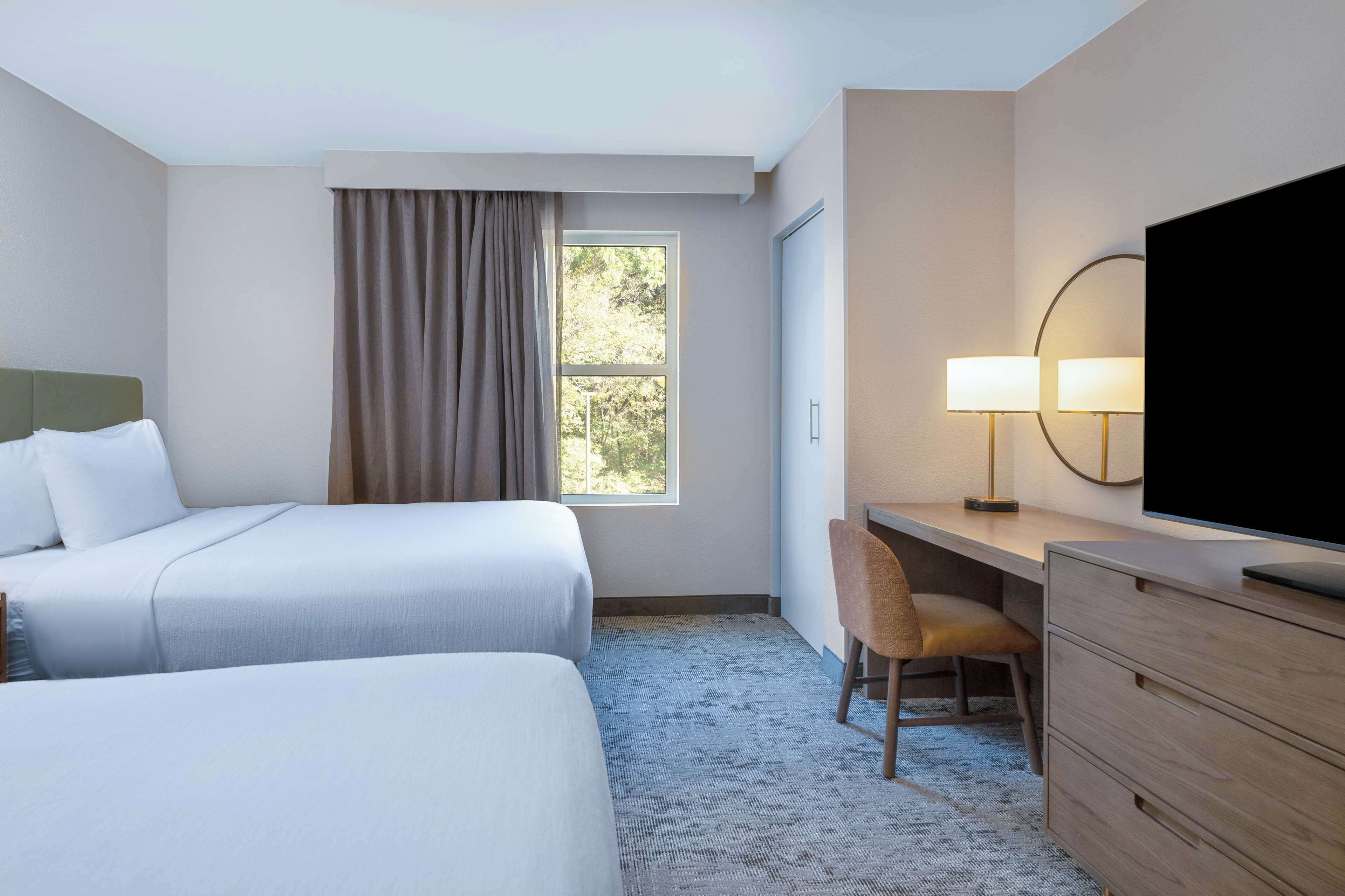 A hotel room with two beds, a desk, a chair, a TV, and a window with curtains.