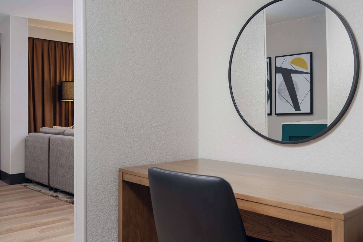 A hotel room with a wooden desk, a black chair, a round mirror, and a framed abstract artwork on the wall.