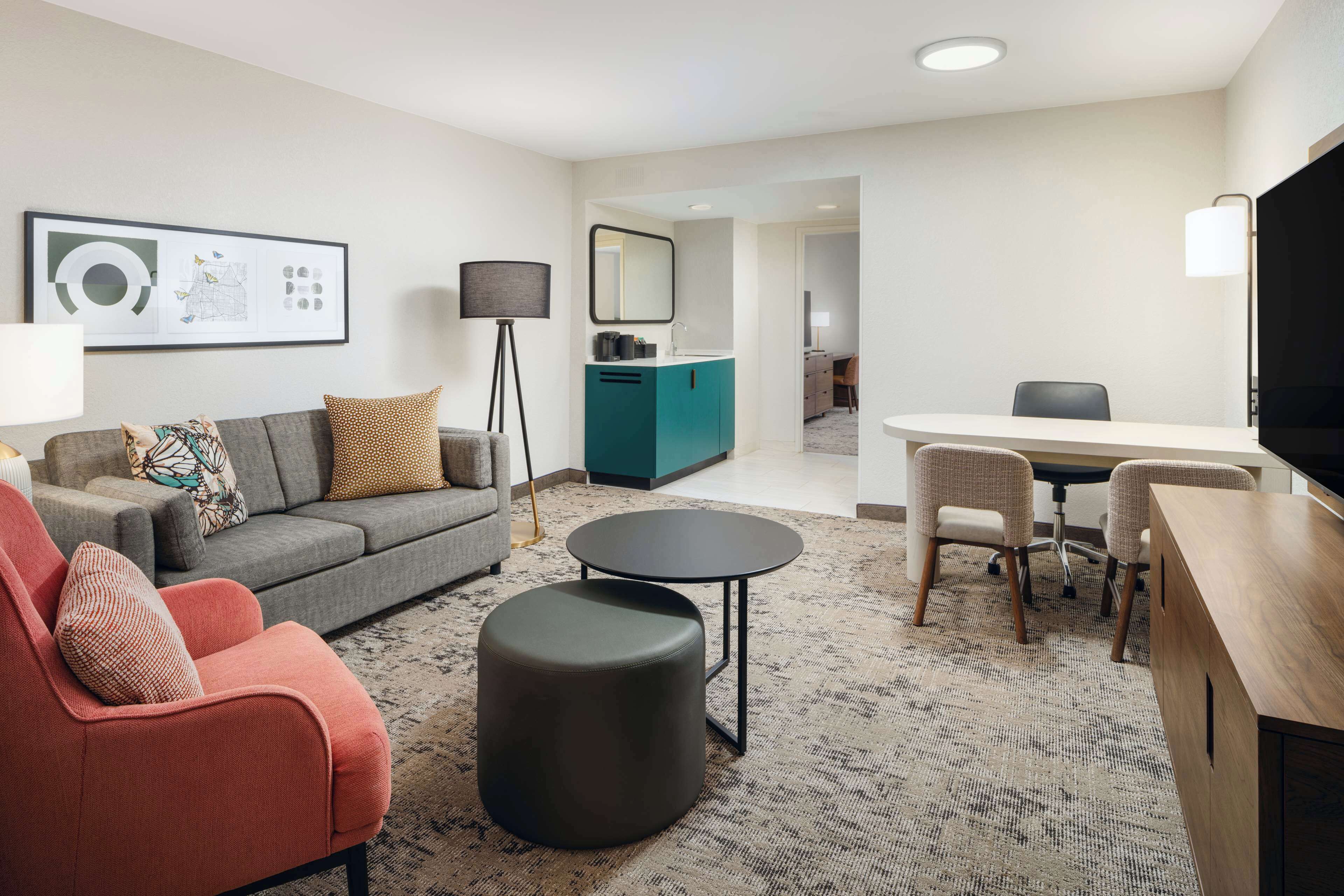 Una sala de estar moderna con un sofá gris, un sillón rojo, un reposapiés negro, una mesa de centro redonda y una zona de comedor con una mesa y sillas blancas.