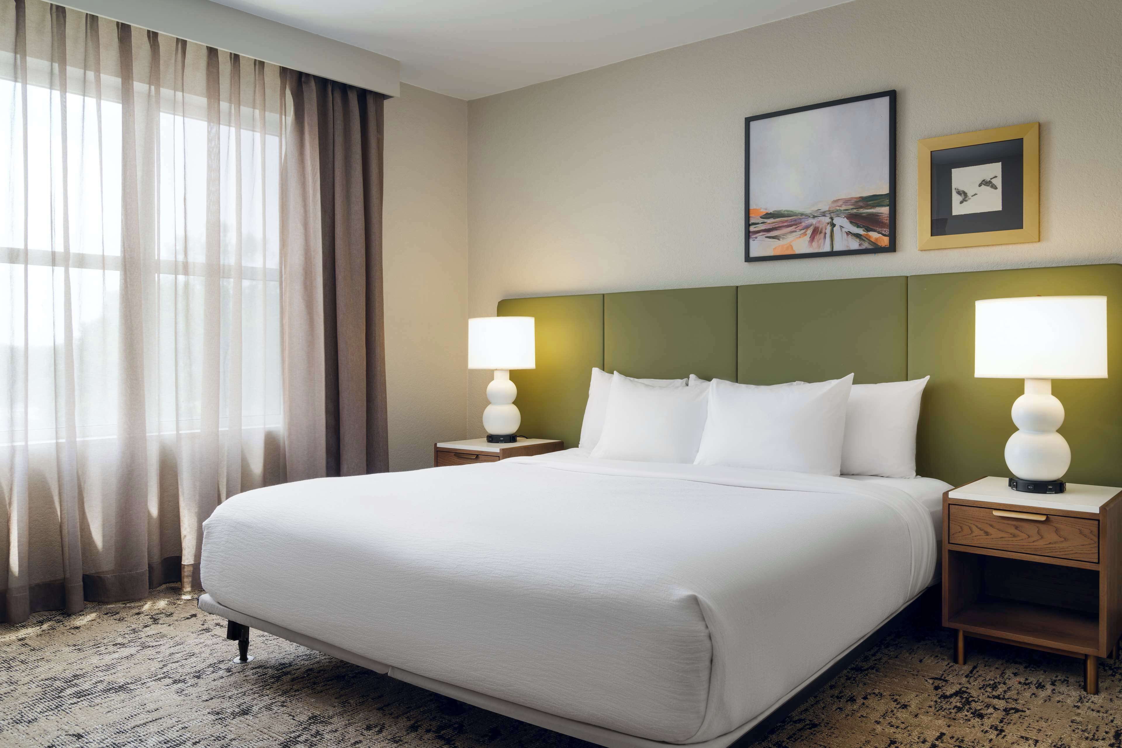 Una habitación de hotel con una cama grande, dos mesitas de noche con lámparas, un cabecero verde y una ventana con cortinas traslúcidas.