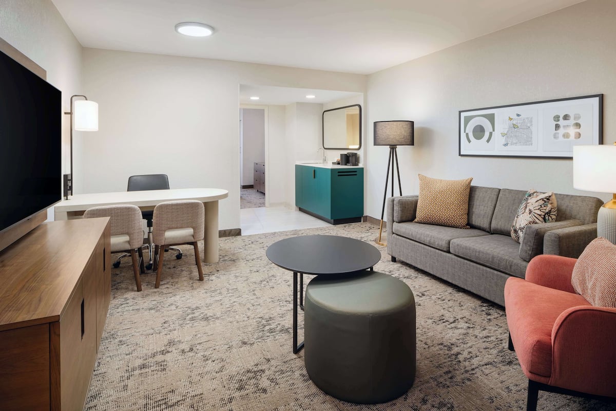 A modern hotel room with a wooden desk, a grey sofa, a round ottoman, a TV, and a framed artwork on the wall.